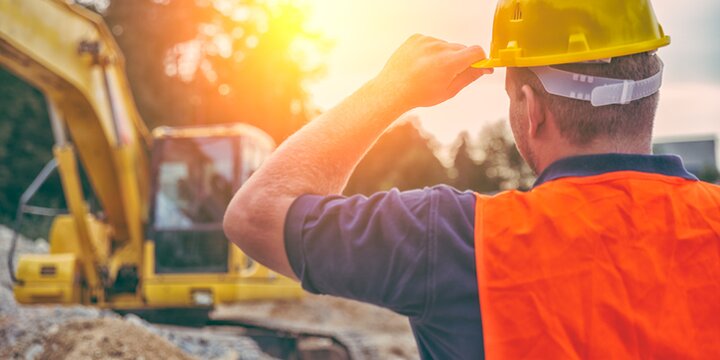 Baggerführer mit Helm mit dem Rücken vor seinem Bagger | © iStock/GregorBister